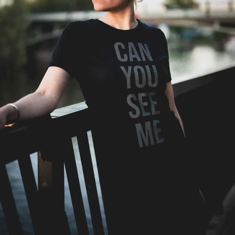 A woman wearing a black T-shirt with the text Can You See Me in black reflective print, standing by a riverside railing._1