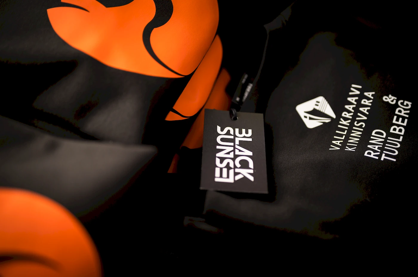 Mustad pusad oranži rebasedisainiga ja Vallikraavi Kinnisvara ning Rand & Tuulberg logodega. Pildil on Blacksunseti eritellimusel valmistatud rõivad, mis sobivad nii firmaüritustele kui ka igapäevaseks kandmiseks.