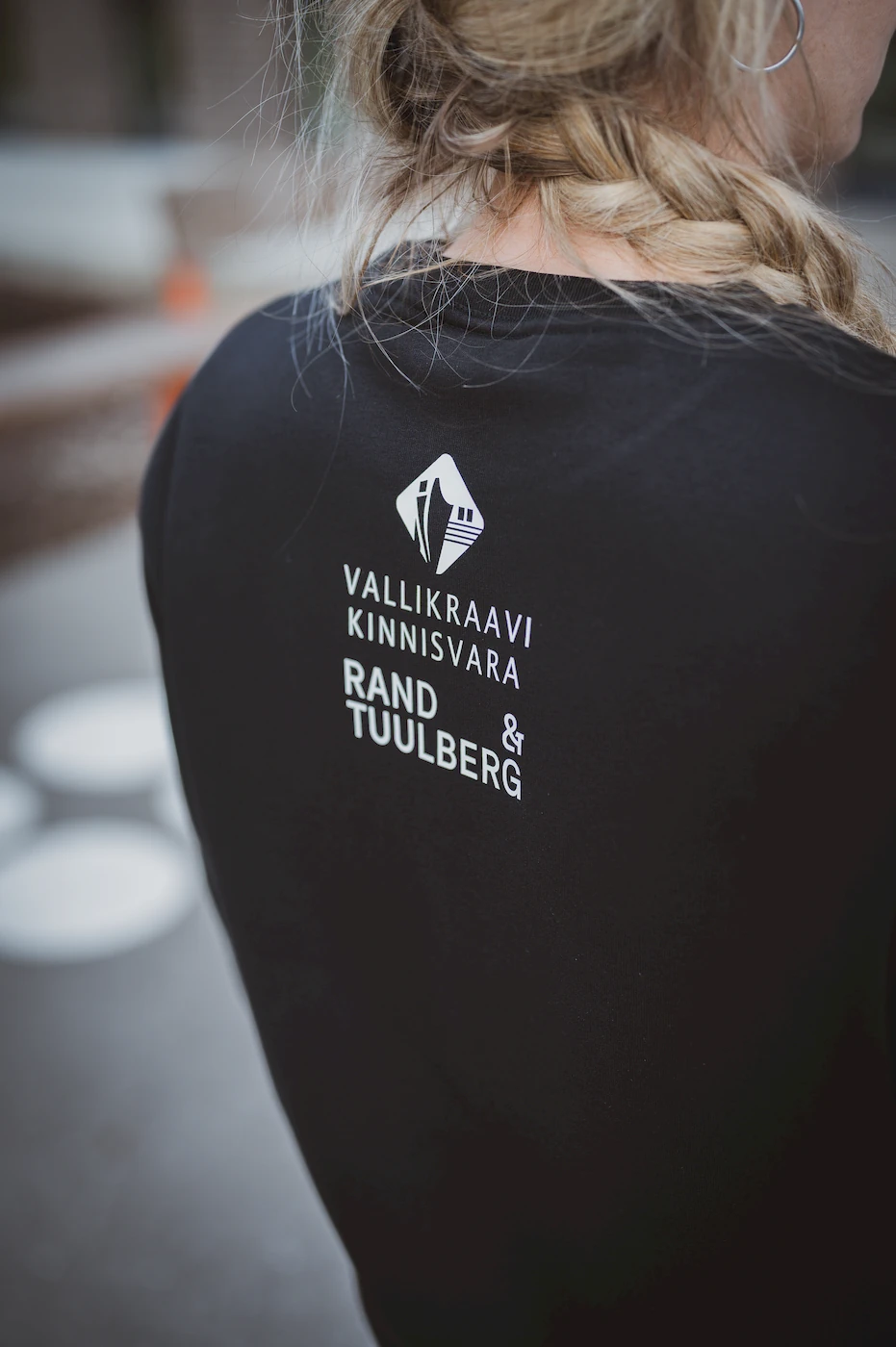 Naine kannab musta pusa, millel on seljal Vallikraavi Kinnisvara ja Rand & Tuulberg logod. Pilt rõhutab ettevõtte brändingut ja professionaalset ilmet, olles osa Blacksunseti eritellimusel valmistatud riiete kollektsioonist.