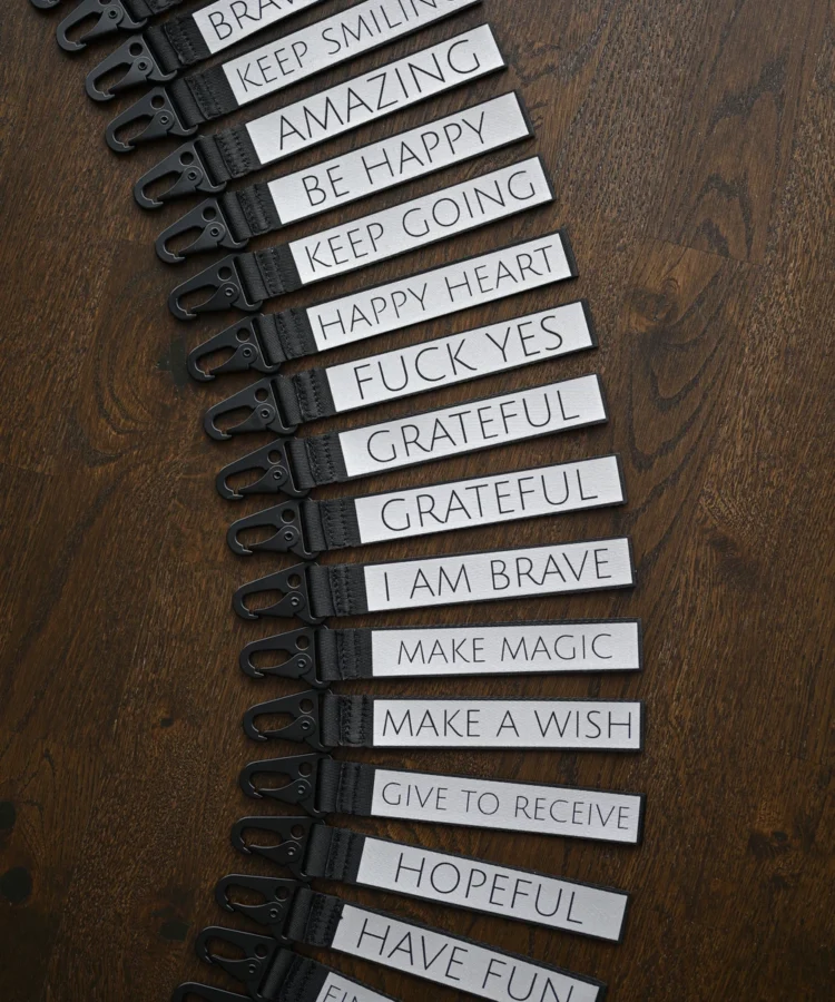 Pikk rida helkurvõtmehoidjaid kaarena; nähtavad tekstid: “GIVE TO RECEIVE”, “MAKE A WISH”, “MAKE MAGIC”, “I AM BRAVE”, “GRATEFUL”, “FUCK YES”, “HAPPY HEART”, “KEEP GOING”, “BE HAPPY”, “AMAZING”, “KEEP SMILING”, “BRAVE SOUL”, “LIFE IS GOOD”, “JUST BREATHE ♥”, “GET LOST”, “FUCK OFF”.
