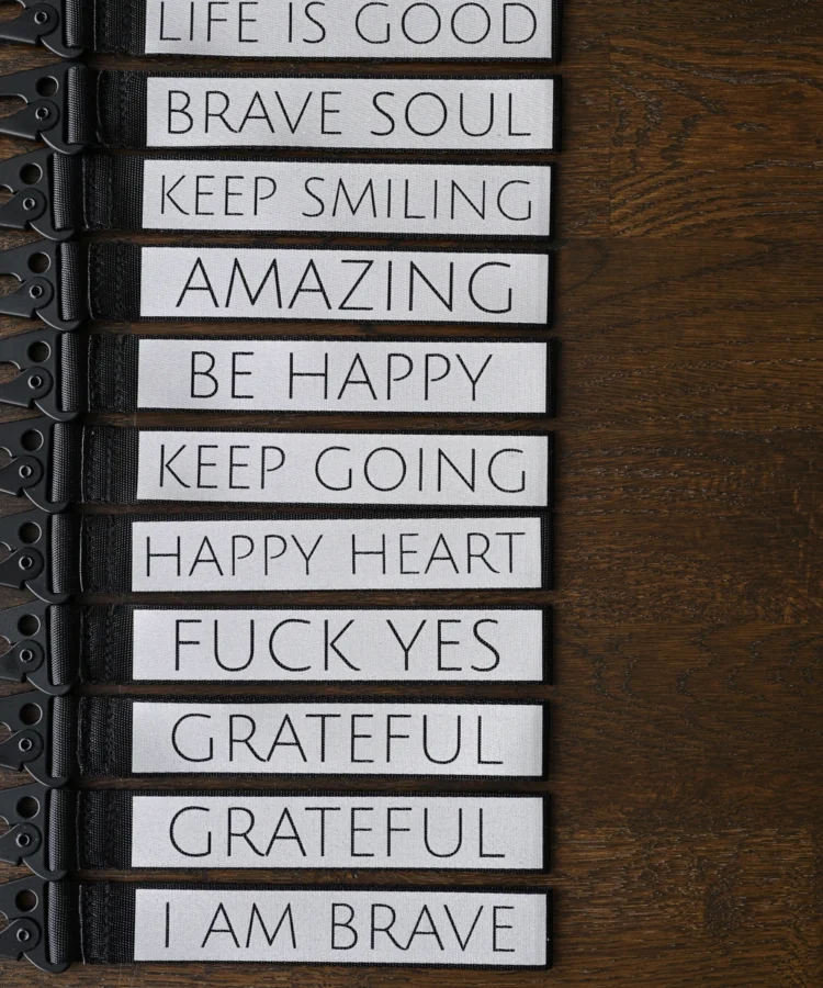 Pikk rida helkurvõtmehoidjaid kaarena; nähtavad tekstid: “GIVE TO RECEIVE”, “MAKE A WISH”, “MAKE MAGIC”, “I AM BRAVE”, “GRATEFUL”, “FUCK YES”, “HAPPY HEART”, “KEEP GOING”, “BE HAPPY”, “AMAZING”, “KEEP SMILING”, “BRAVE SOUL”, “LIFE IS GOOD”, “JUST BREATHE ♥”, “GET LOST”, “FUCK OFF”.