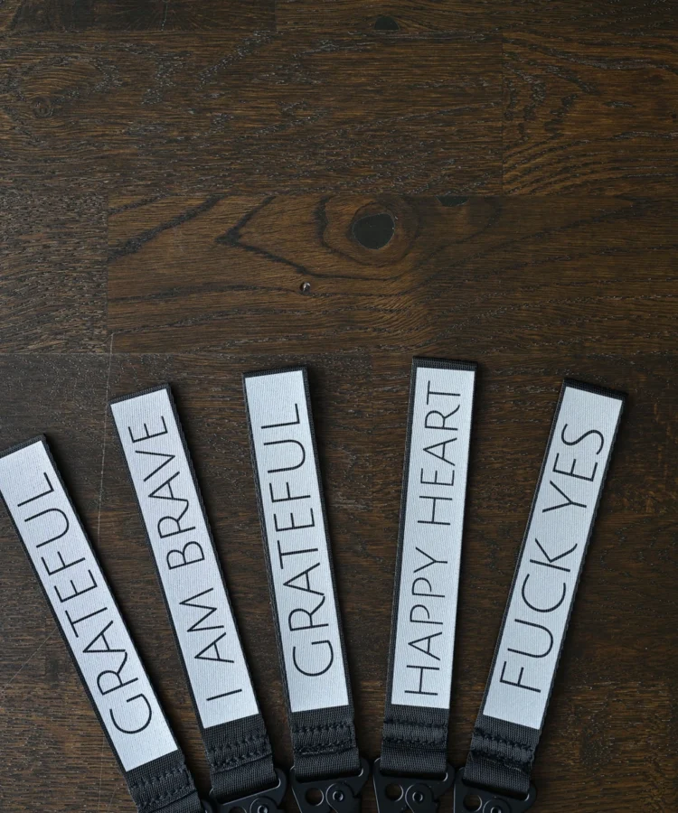 Viis helkurvõtmehoidjat puidust taustal lehvikuna; tekstid: “GRATEFUL”, “I AM BRAVE”, “GRATEFUL”, “HAPPY HEART”, “FUCK YES”.