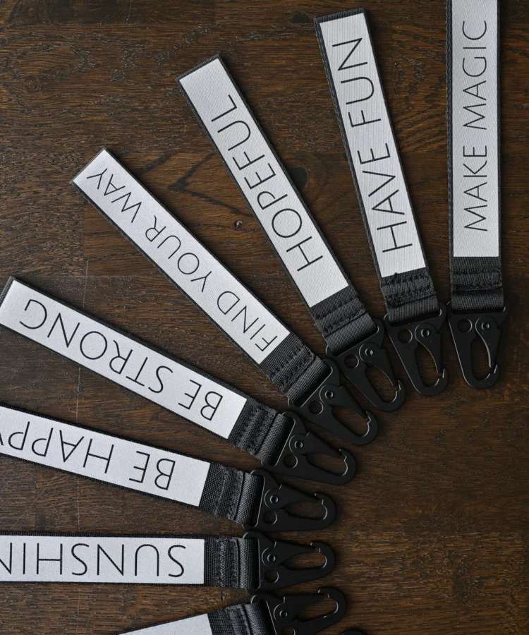 Helkurvõtmehoidjate valik lehvikuna, mustad karabiinid; nähtavad tekstid: “SMILE :)”, “SUNSHINE”, “BE HAPPY”, “BE STRONG”, “FIND YOUR WAY”, “HOPEFUL”, “HAVE FUN”, “MAKE MAGIC”.