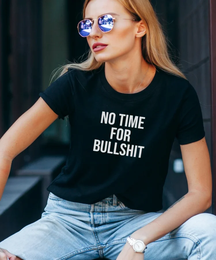 Naine mustas “NO TIME FOR BULLSHIT” T-särgis, päikeseprillid, istub linnakeskkonnas; lifestyle-foto.
