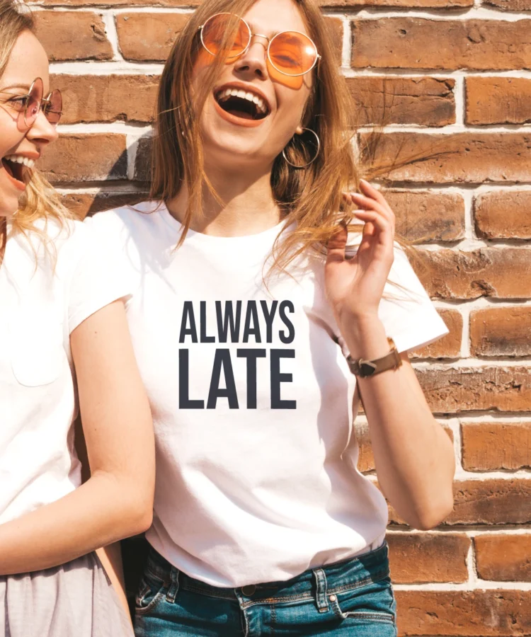 Naeratav naine valges “ALWAYS LATE” T-särgis, koos sõbrannaga, lifestyle-foto, tellissein taustal.
