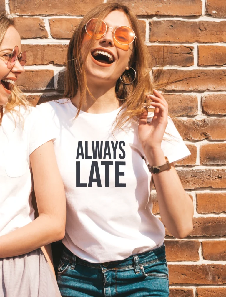 Naeratav naine valges “ALWAYS LATE” T-särgis, koos sõbrannaga, lifestyle-foto, tellissein taustal.