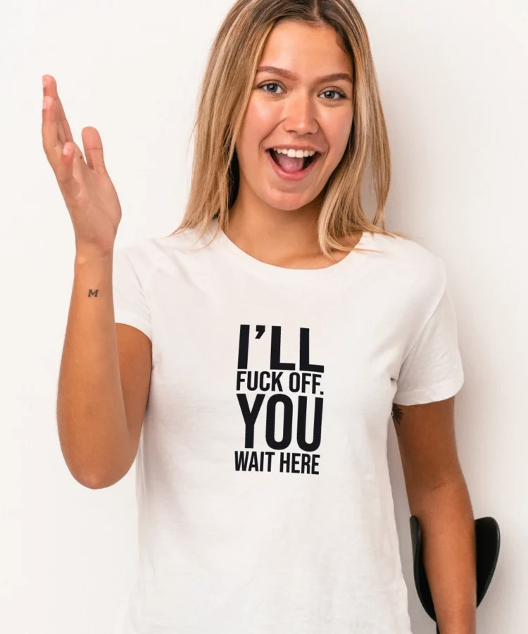 Naeratav naine valges “I’LL FUCK OFF. YOU WAIT HERE” T-särgis, seisab heleda taustaga; lifestyle-foto.