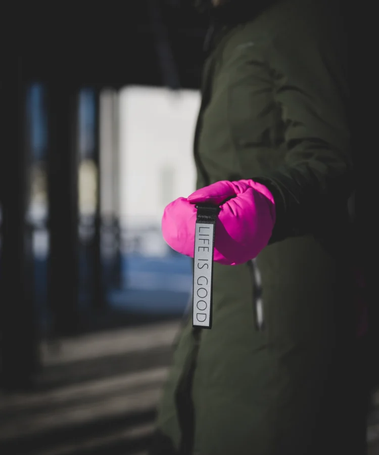 Roosa kinnas hoiab horisontaalselt helkurvõtmehoidjat tekstiga “LIFE IS GOOD”; talvine väliskeskkond, jope ja varrukas fookusest väljas.