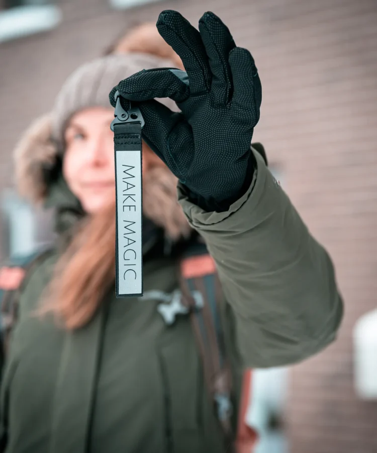 Inimene mustas kindas hoiab vertikaalselt helkurvõtmehoidjat tekstiga “MAKE MAGIC”, taustal tellissein ja talvejope.