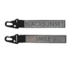 Helkurvõtmehoidja halli peegeldava ribaga ja musta karabiiniga; ühelt poolt tekst “BLACKSUNSET”, teiselt “SMILE :)”, tootekujutis valgel taustal.