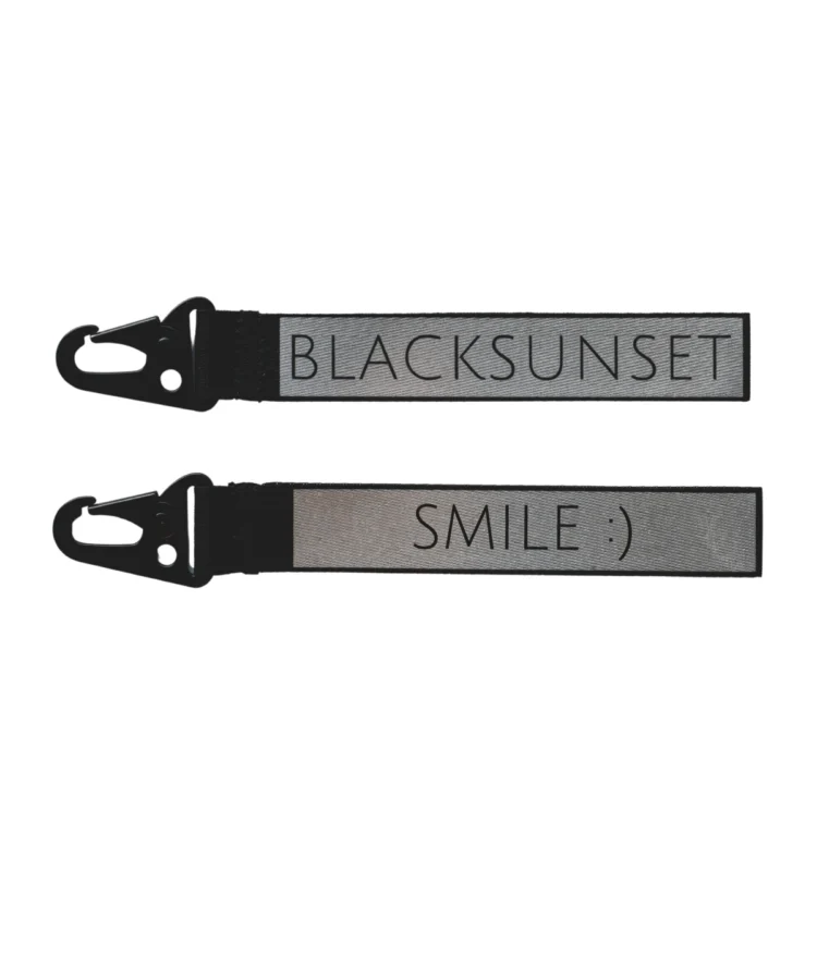Helkurvõtmehoidja halli peegeldava ribaga ja musta karabiiniga; ühelt poolt tekst “BLACKSUNSET”, teiselt “SMILE :)”, tootekujutis valgel taustal.