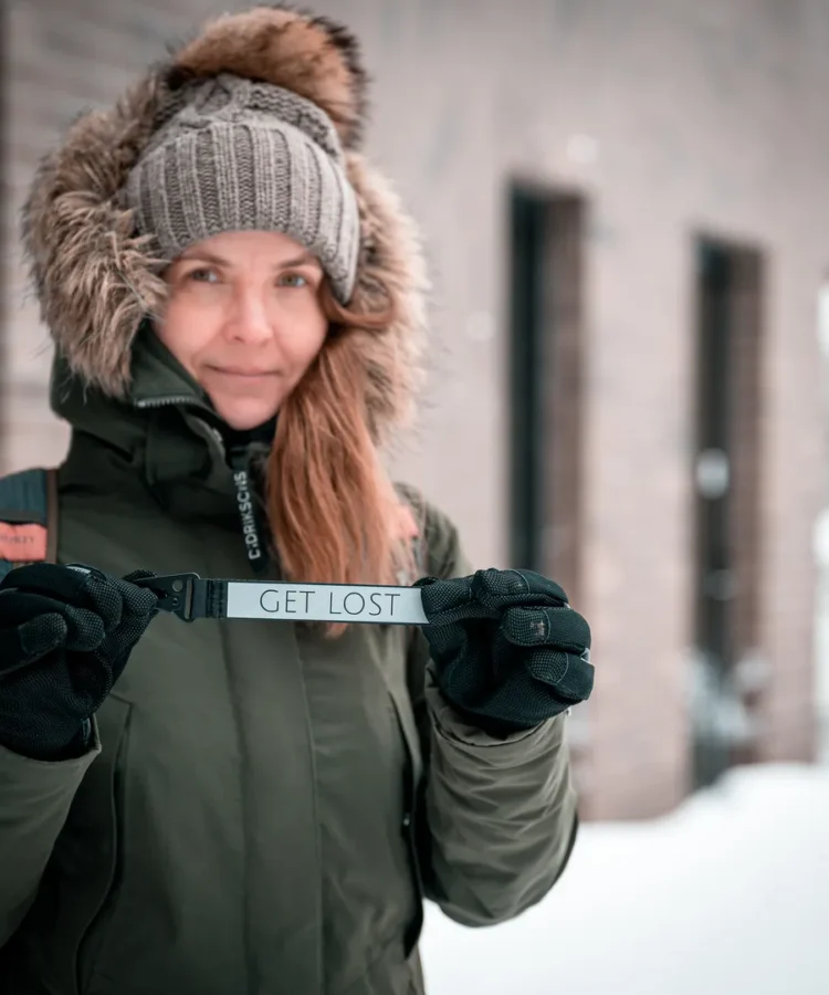 Inimene hoiab kahel käel helkurvõtmehoidjat tekstiga “GET LOST”; talveriided, taustal hoone ja lumi.