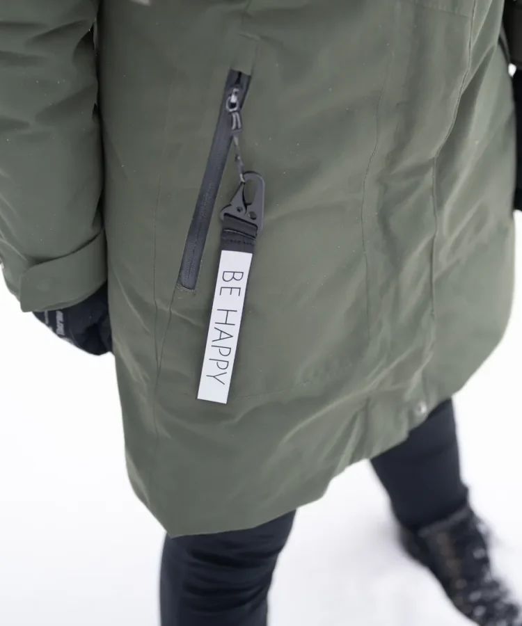 Helkurvõtmehoidja tekstiga “BE HAPPY” ripub talvejope lukul; lumi maas, rõhk tootel.