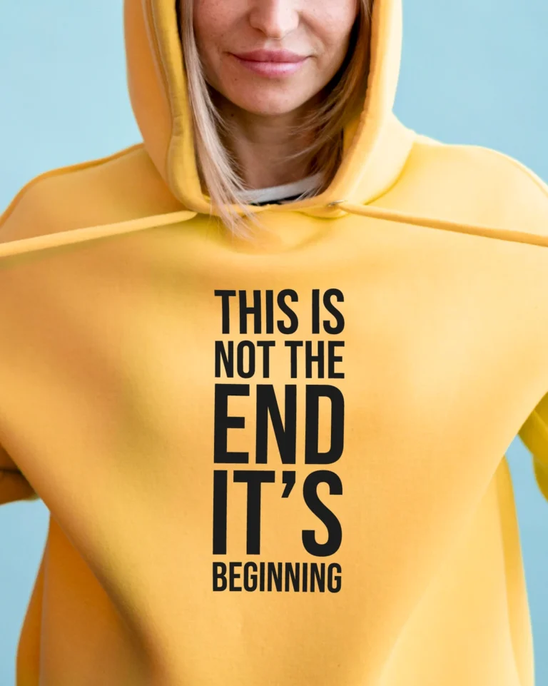 Kollane kapuutsikas tekstiga “THIS IS NOT THE END IT’S BEGINNING” – suur must front-print; stuudiopilt, lifestyle-visuaal.