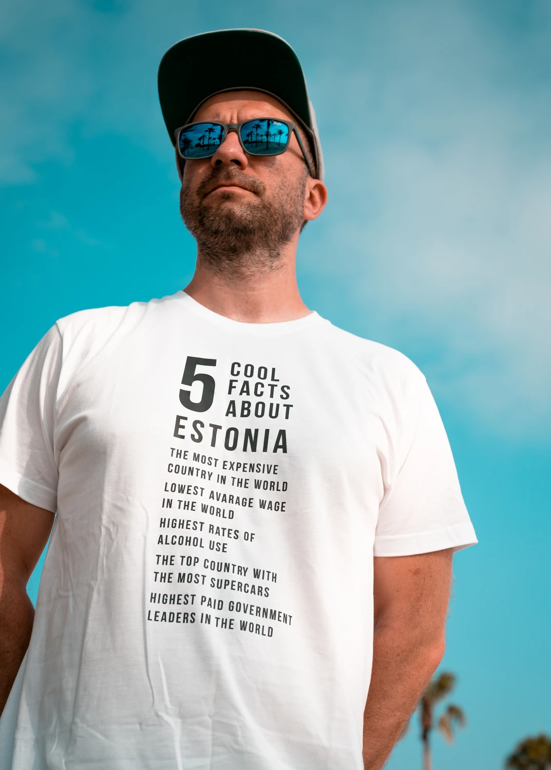 Mees kannab valget Blacksunset “5 Cool Facts About Estonia” T-särki; päikeseprillid ja nokkmüts, sinine taevas taustal.