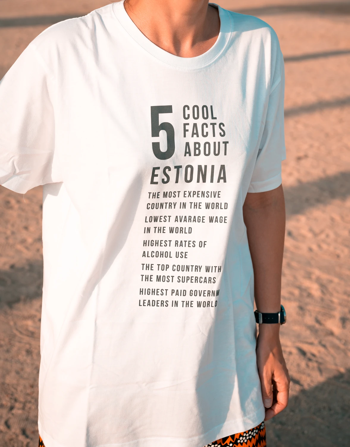 Valge Blacksunset “5 Cool Facts About Estonia” T-särk modellil rannaliival; vertikaalne tekstidisain.