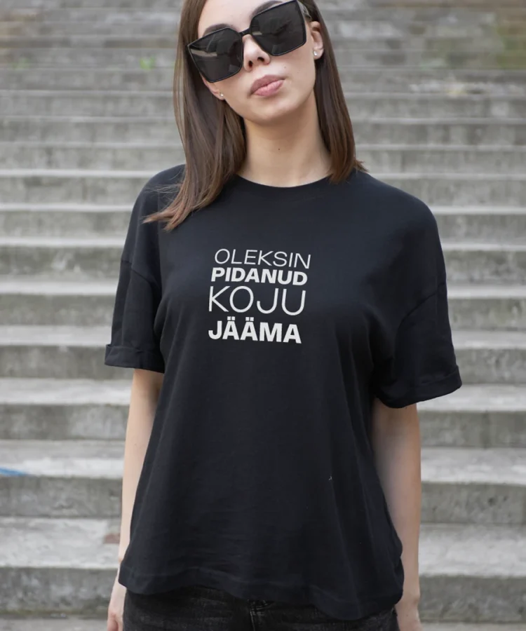 Noor naine suurte mustade päikeseprillidega trepiastmetel; seljas must BLACKSUNSET T-särk valge kirjaga “OLEKSIN PIDANUD KOJU JÄÄMA” – urban/street-stiilis lifestyle foto.