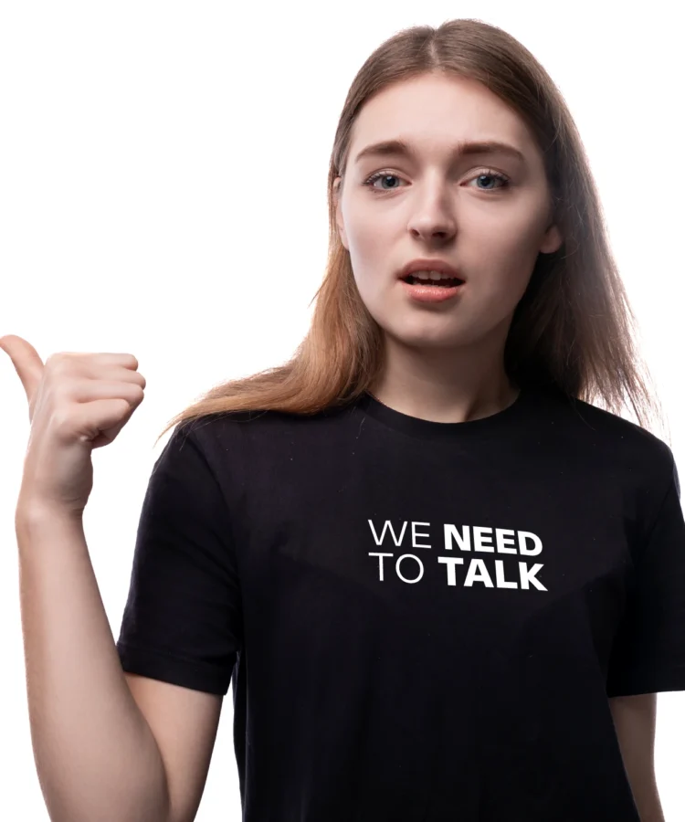 Noor naine musta “WE NEED TO TALK” T-särgiga, osutab pöidlaga vasakule; stuudiotaust, mudelifoto.