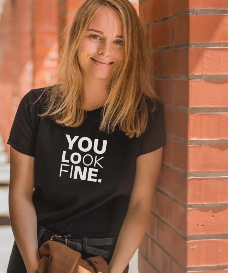 oor naine kannab musta Blacksunset unisex T-särki kirjaga “YOU LOOK FINE.” – lifestyle/modellifoto punase tellisseina juures.