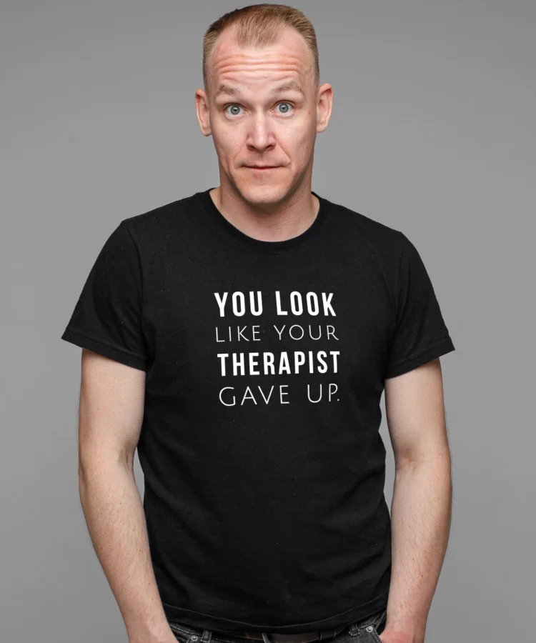 Mees stuudios mustas T-särgis valge tekstiga “YOU LOOK LIKE YOUR THERAPIST GAVE UP.”; hall taust, käed taskus, otsevaates.