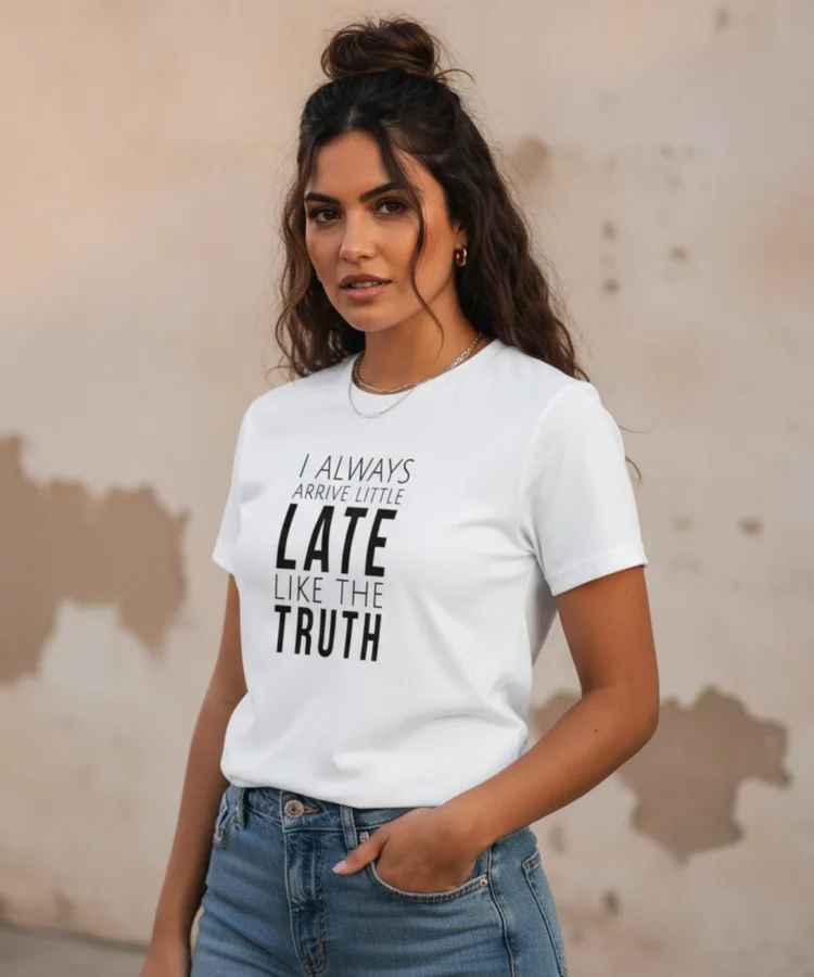 Blacksunset unisex valge T-särk „I ALWAYS ARRIVE LITTLE LATE LIKE THE TRUTH”; ¾ eesvaade, käsi taskus, kulunud beež seintaust, soe õhtuvalgus.