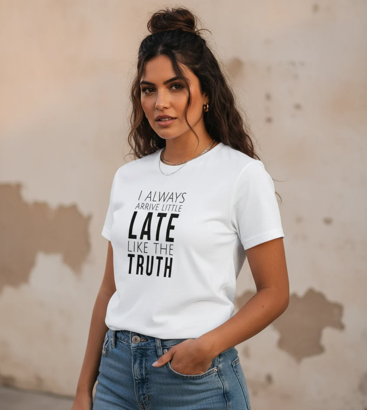 Blacksunset unisex valge T-särk „I ALWAYS ARRIVE LITTLE LATE LIKE THE TRUTH”; ¾ eesvaade, käsi taskus, kulunud beež seintaust, soe õhtuvalgus.