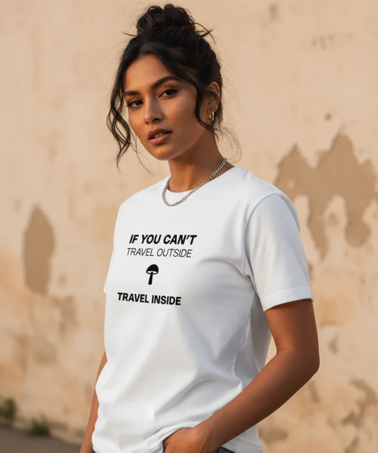 Blacksunset valge unisex T-särk tekstiga „IF YOU CAN’T TRAVEL OUTSIDE / TRAVEL INSIDE”; must kiri ja seen, naismudel ¾ vaates, kulunud seina taust.