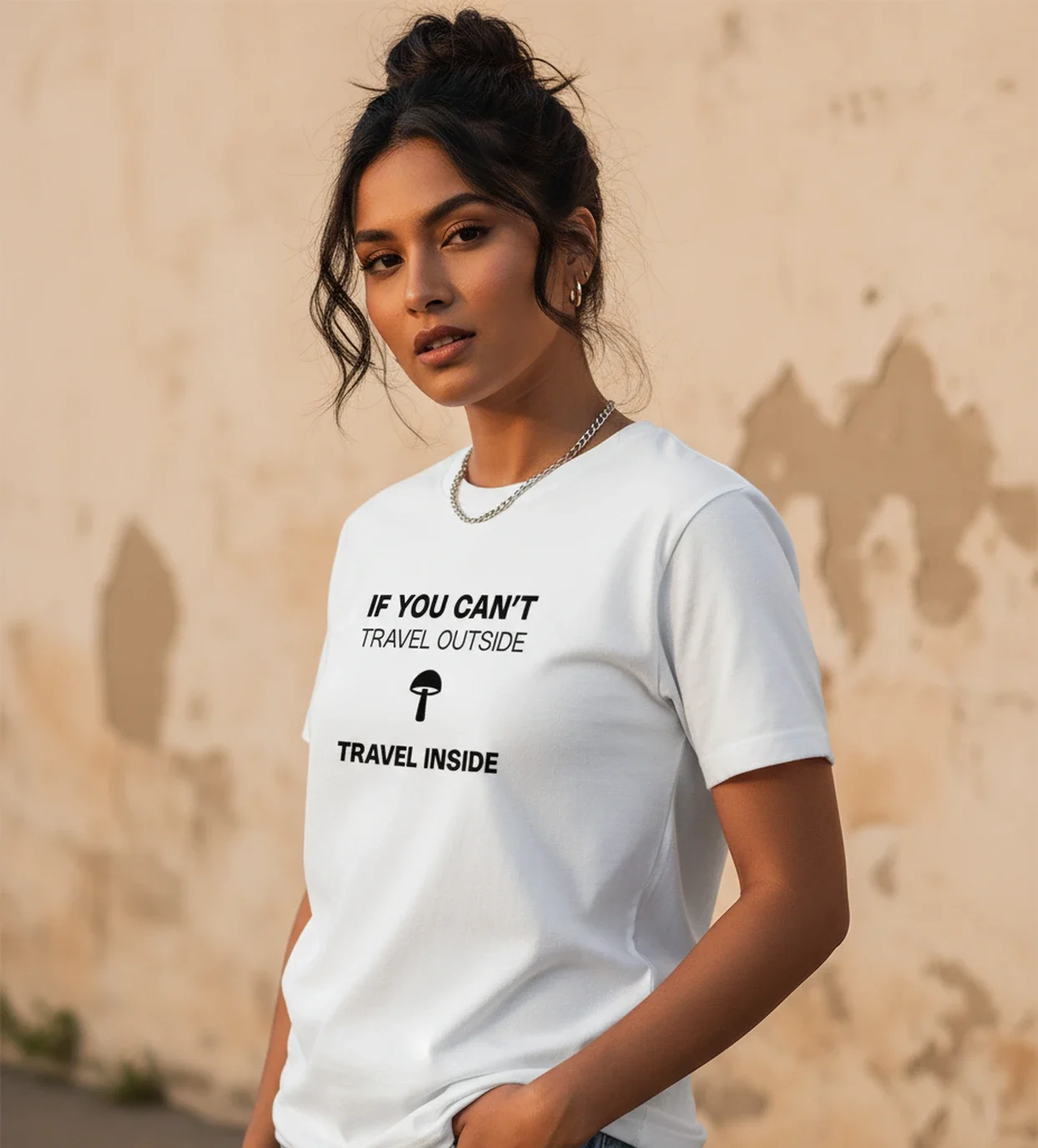 Blacksunset valge unisex T-särk tekstiga „IF YOU CAN’T TRAVEL OUTSIDE / TRAVEL INSIDE”; must kiri ja seen, naismudel ¾ vaates, kulunud seina taust.