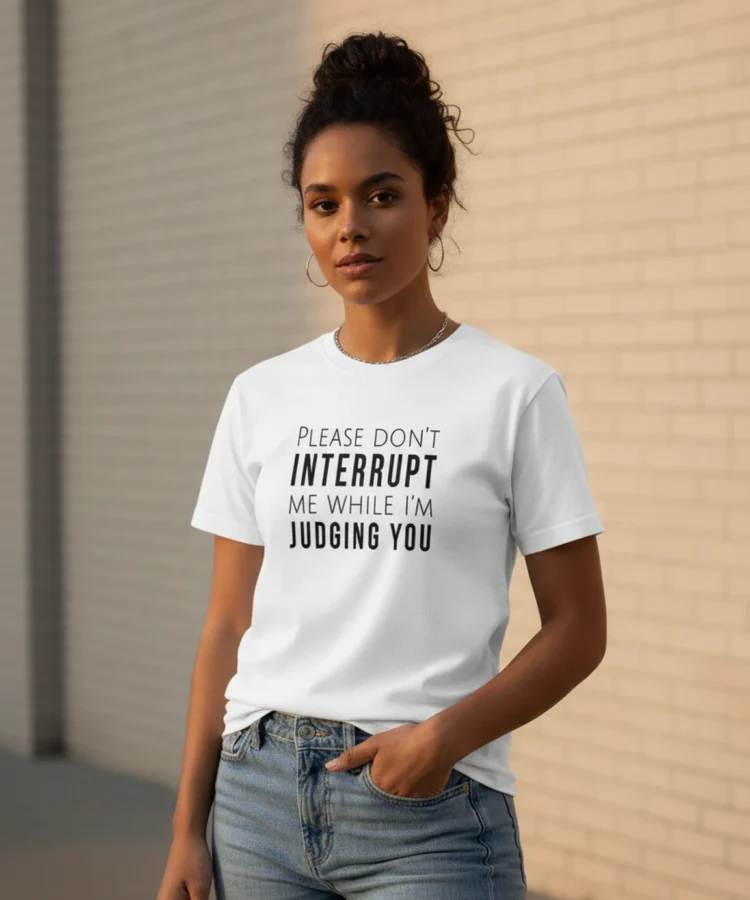 Blacksunset unisex valge T-särk PLEASE DON'T INTERRUPT ME WHILE I'M JUDGING YOU — must kiri; 3/4 profiil, käsi taskus, soe õhtuvalgus, tellissein.