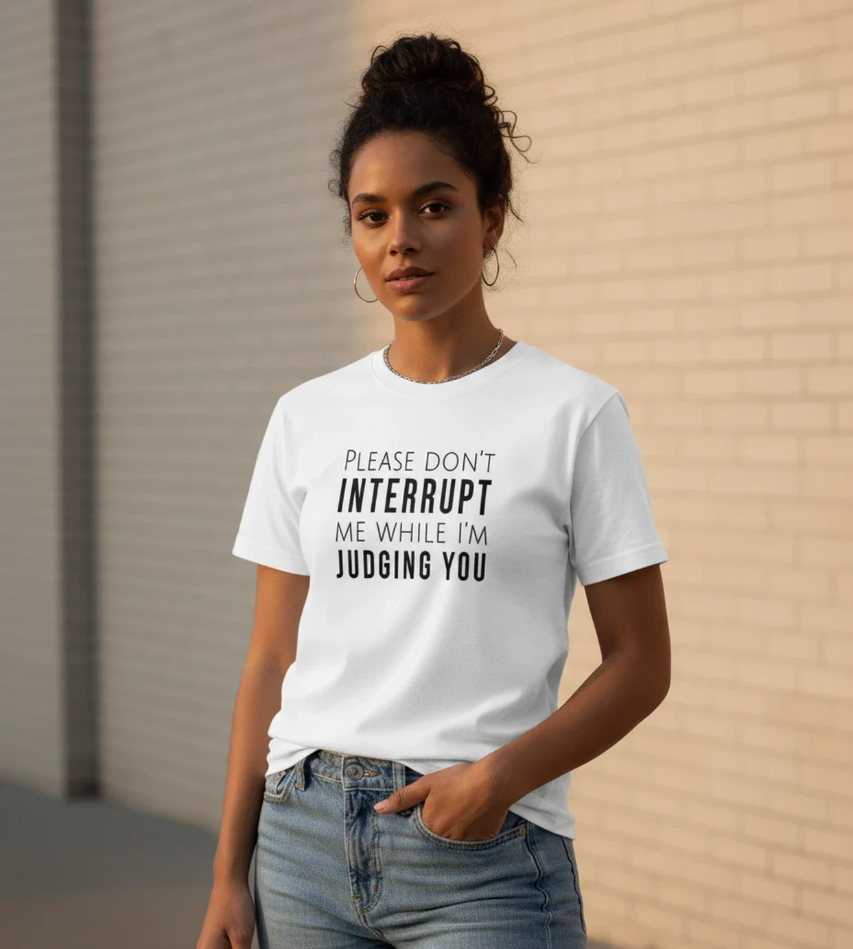 Blacksunset unisex valge T-särk PLEASE DON'T INTERRUPT ME WHILE I'M JUDGING YOU — must kiri; 3/4 profiil, käsi taskus, soe õhtuvalgus, tellissein.
