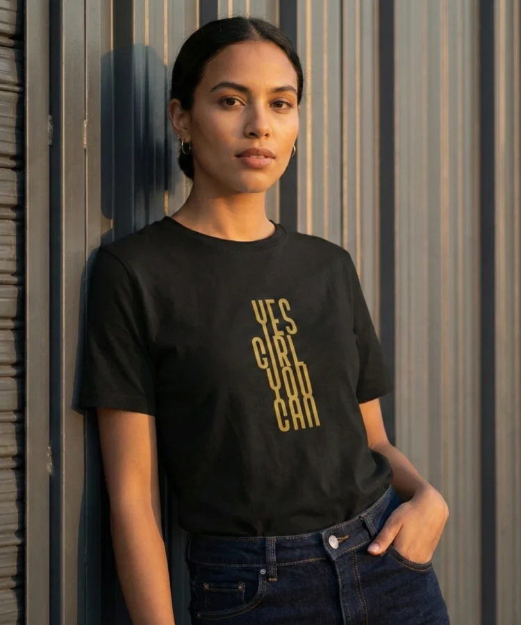 Modell mustas Blacksunset unisex T-särgis “YES GIRL YOU CAN”, kuldne vertikaalne tekst, metallseina taustal.