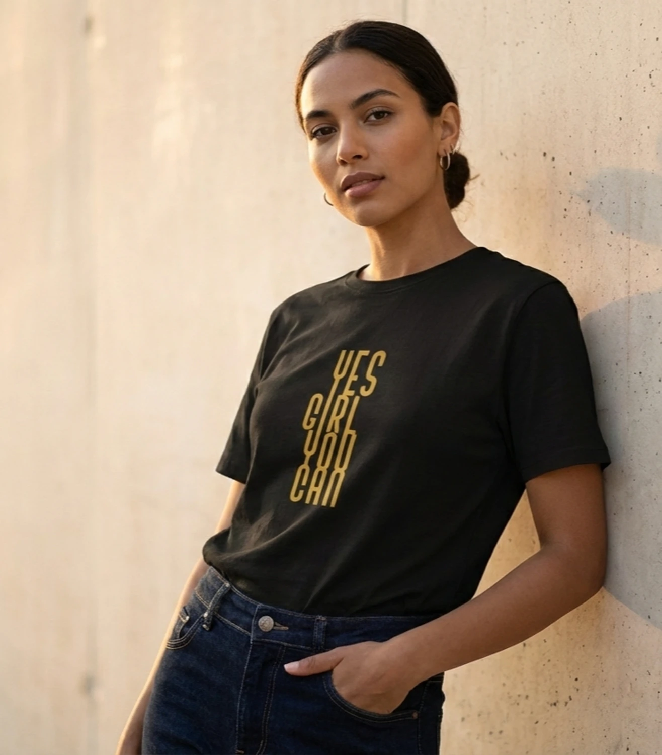 Modell kannab musta Blacksunset unisex T-särki “YES GIRL YOU CAN” kuldse vertikaalse trükiga, betoonseina taustal.