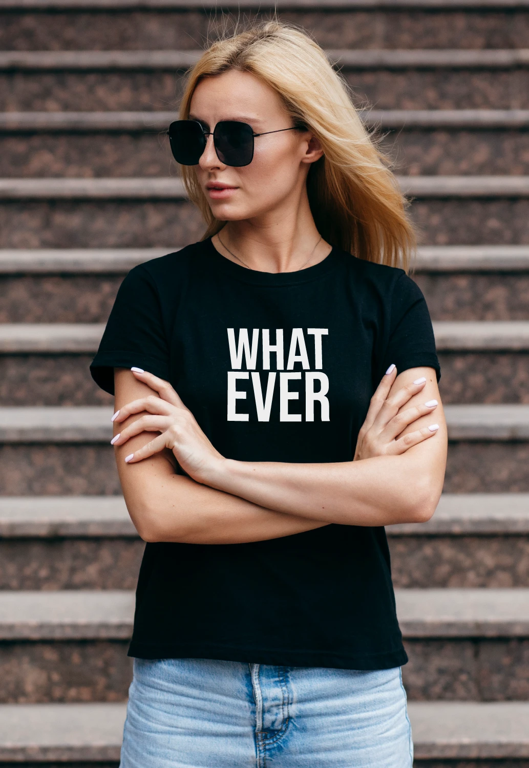 A woman wearing a black WHAT EVER T-shirt and sunglasses, standing with her arms crossed on steps.