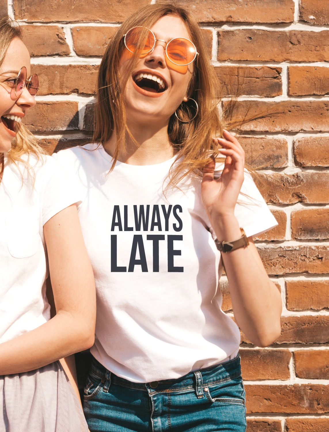 A smiling woman wearing a white ALWAYS LATE T-shirt with a friend, against a brick wall.