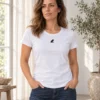Carolina wears a white fitted women's Kapibaara T-shirt and blue jeans in a bright garden room, with a small black logo at the centre of her chest.