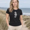 Liis wears the black women's-cut Merihobu T-shirt on a coastal path. The white seahorse print is fully visible on the front.
