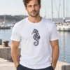 Rasmus wears the white unisex Merihobu T-shirt at a quiet harbour. A black ornamental seahorse is centred on the front.