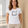 Carolina wears the white women's-cut Raamist väljas T-shirt beside light wood panels in a bright gallery. A black square frame is centred on the shirt with a dot above and to the right.